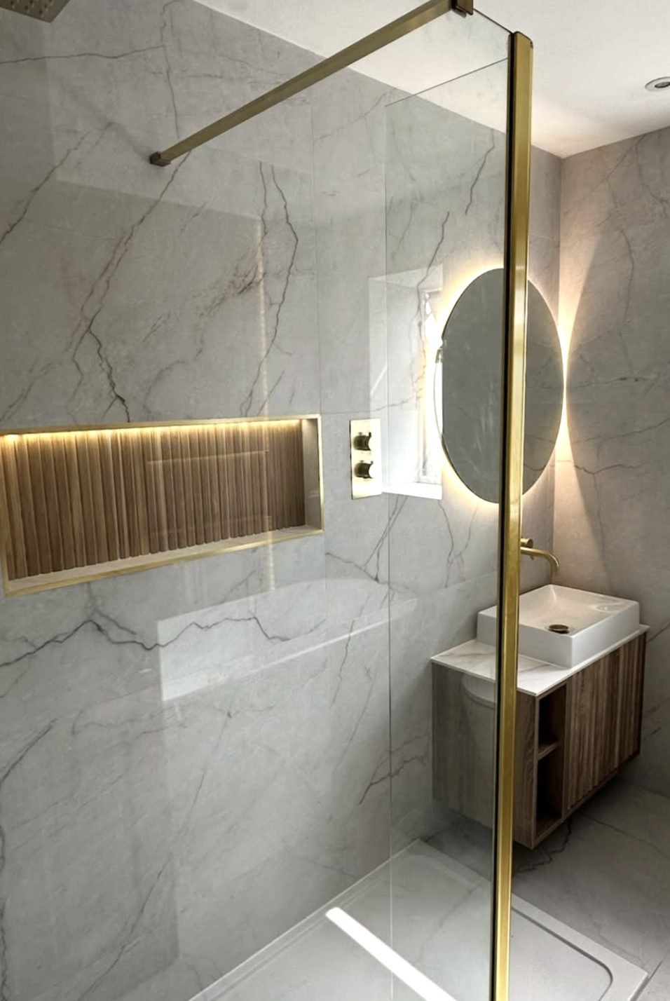 Gold framed shower screen beside a lit fluted recess in a marble wall