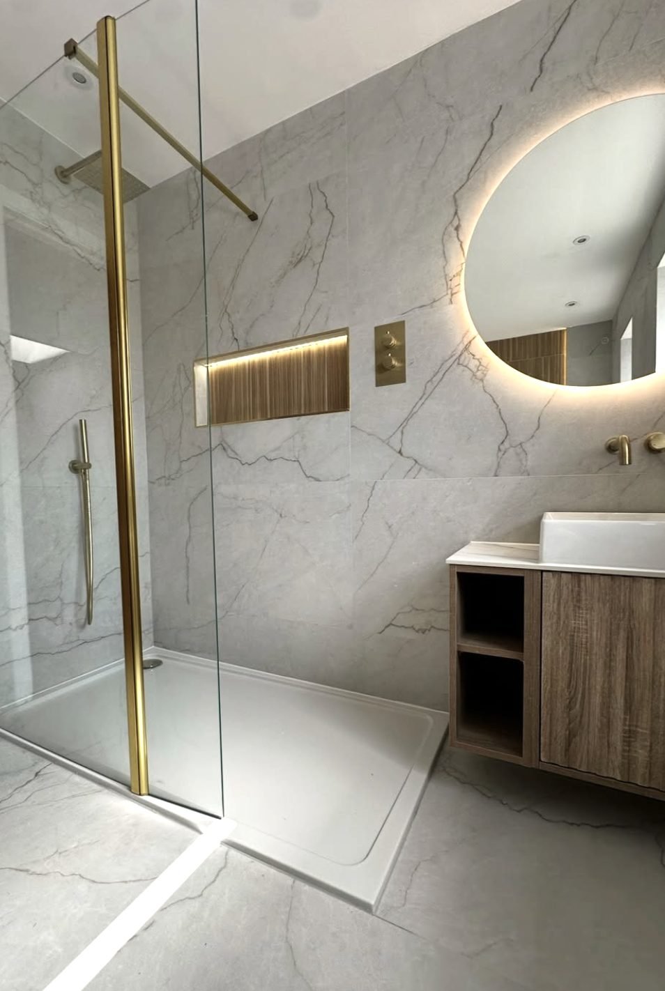 Marble and antique brass walk-in shower with oak veneer vanity and lit round mirror