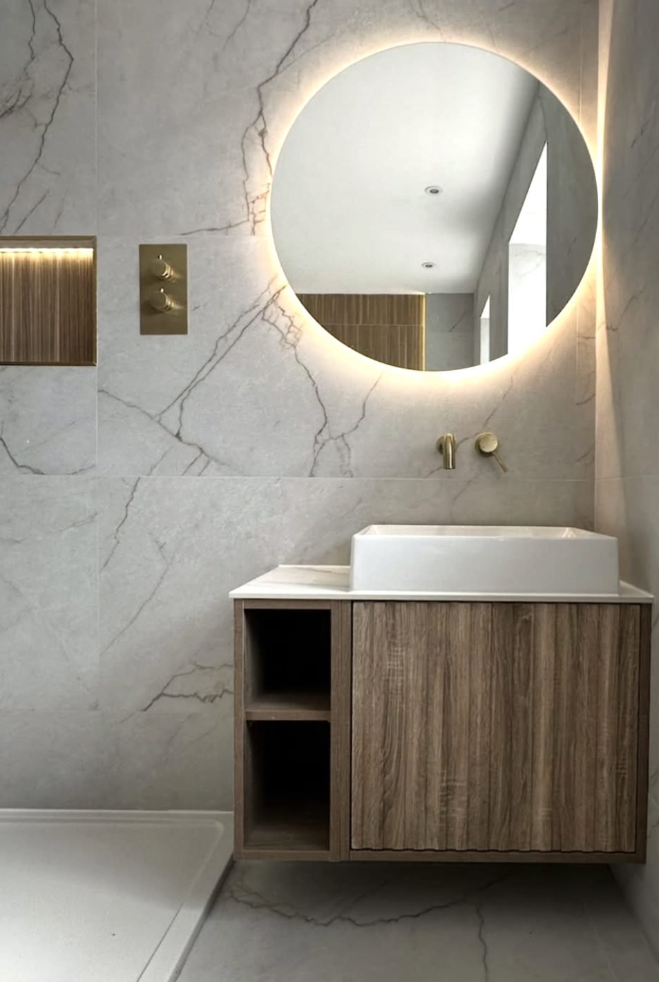 Backlit round mirror above an oak vanity with brass wall-mounted taps