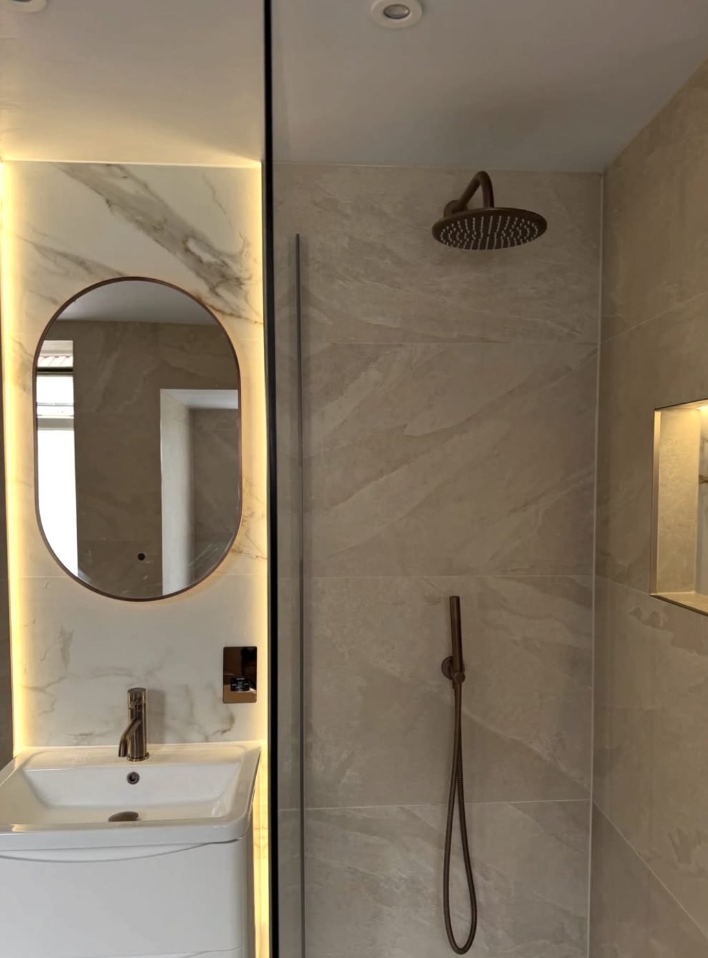 Stone effect shower enclosure with brass rainhead and a backlit oval mirror