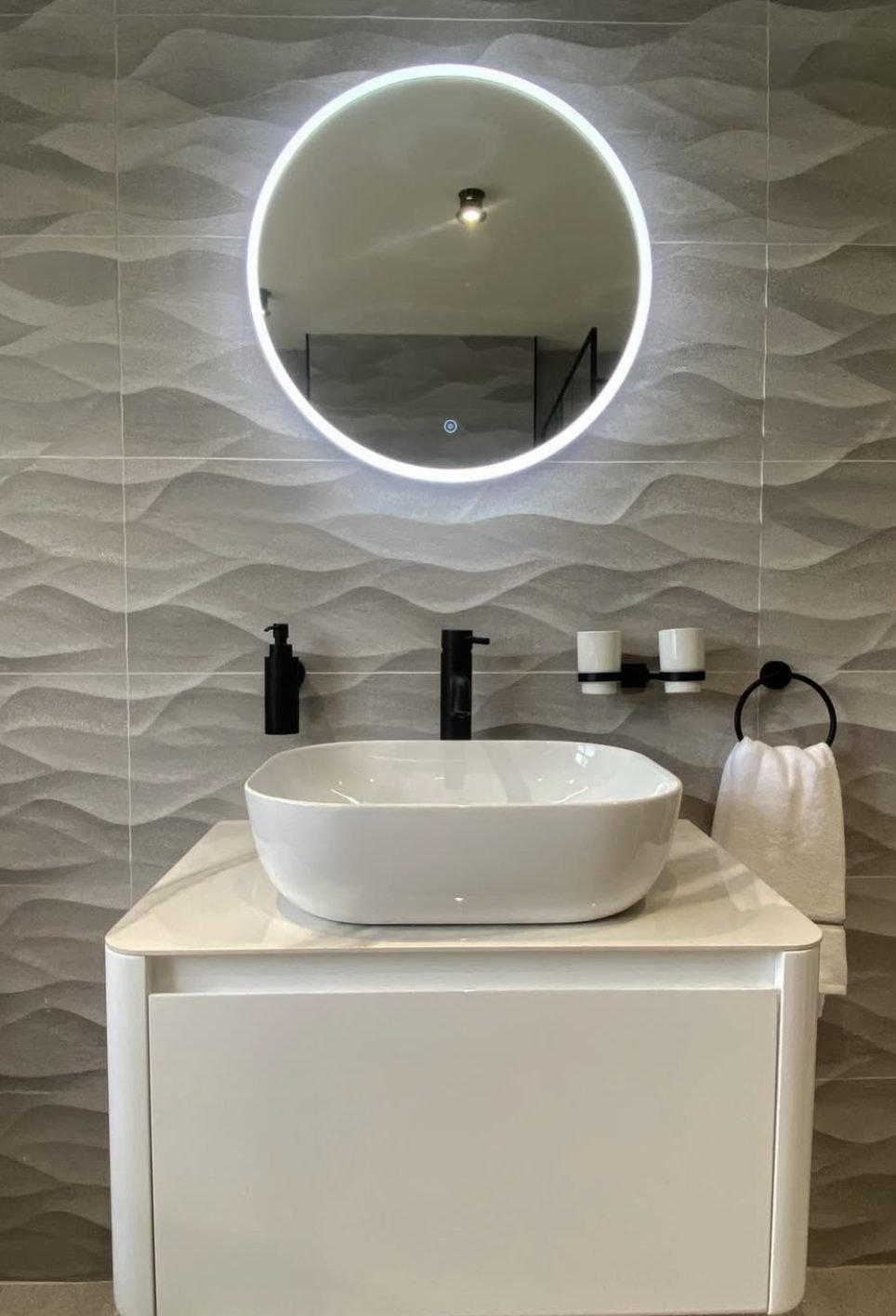 Textured wave tile wall with black fittings, counter basin and round lit mirror